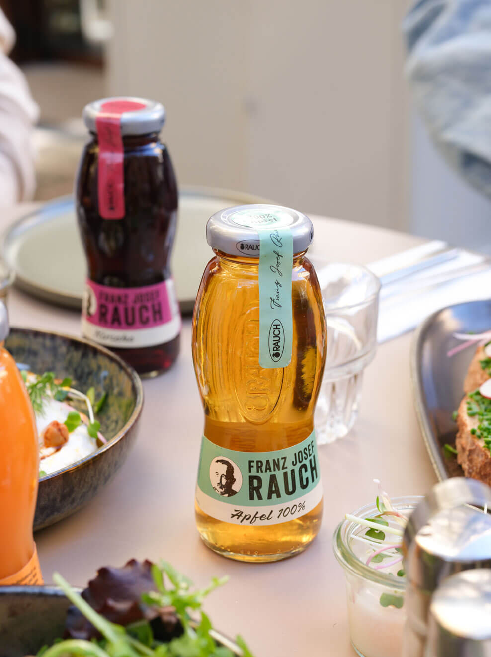 Eine Flasche Franz Josef Rauch 100% Apfelsaft steht auf einem mit Lebensmitteln gedeckten Tisch. Im Hintergrund sind eine weitere Flasche mit einem rosafarbenen Etikett und verschiedene Speisen zu sehen.