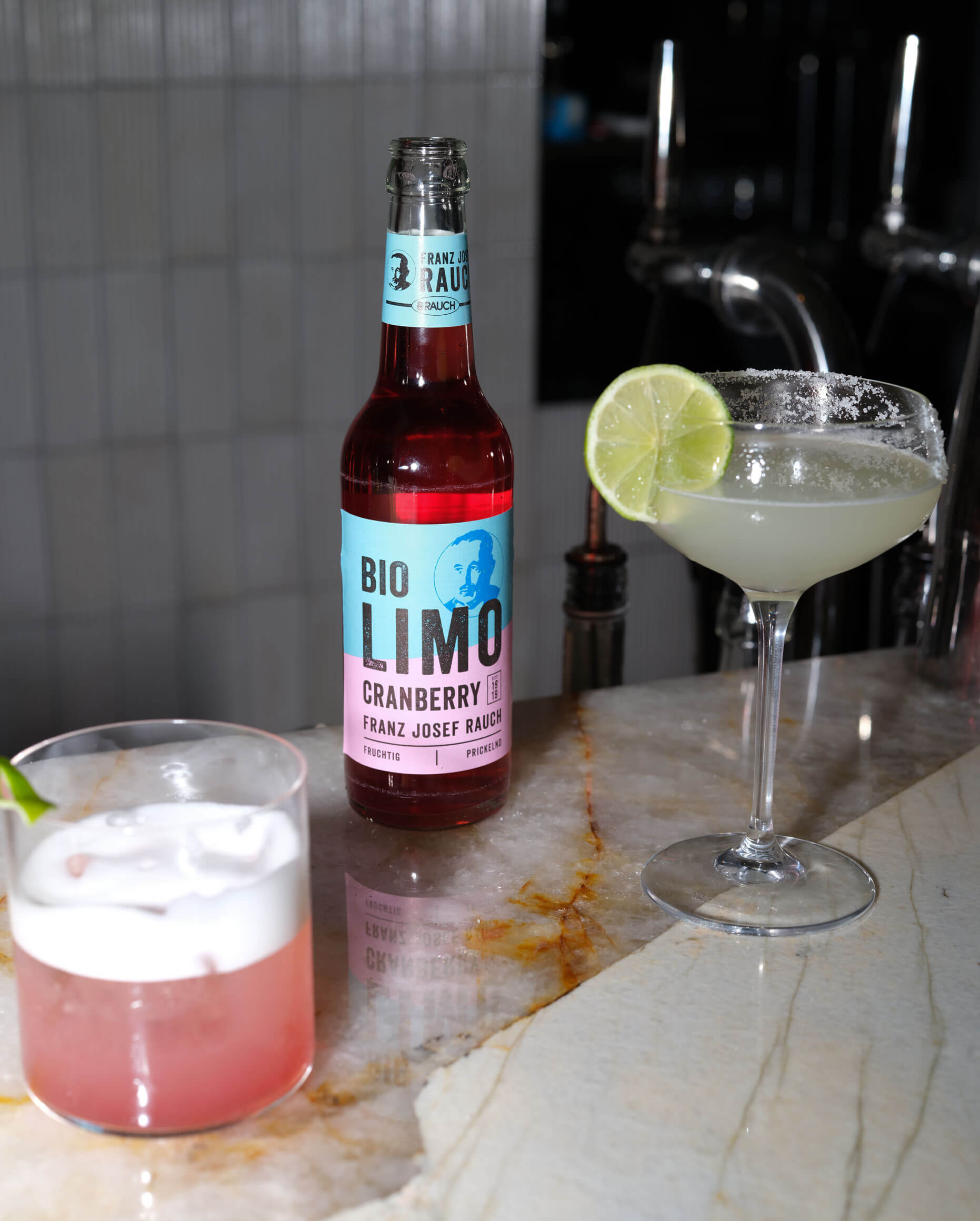 Eine Flasche Franz Josef Rauch Bio Limo Cranberry Soda steht auf einem Marmortresen neben einem Cocktailglas mit Salzrand und Limettenscheibe und einem rosafarbenen Getränk in einem kurzen Glas mit Eis und Limettenspalte.