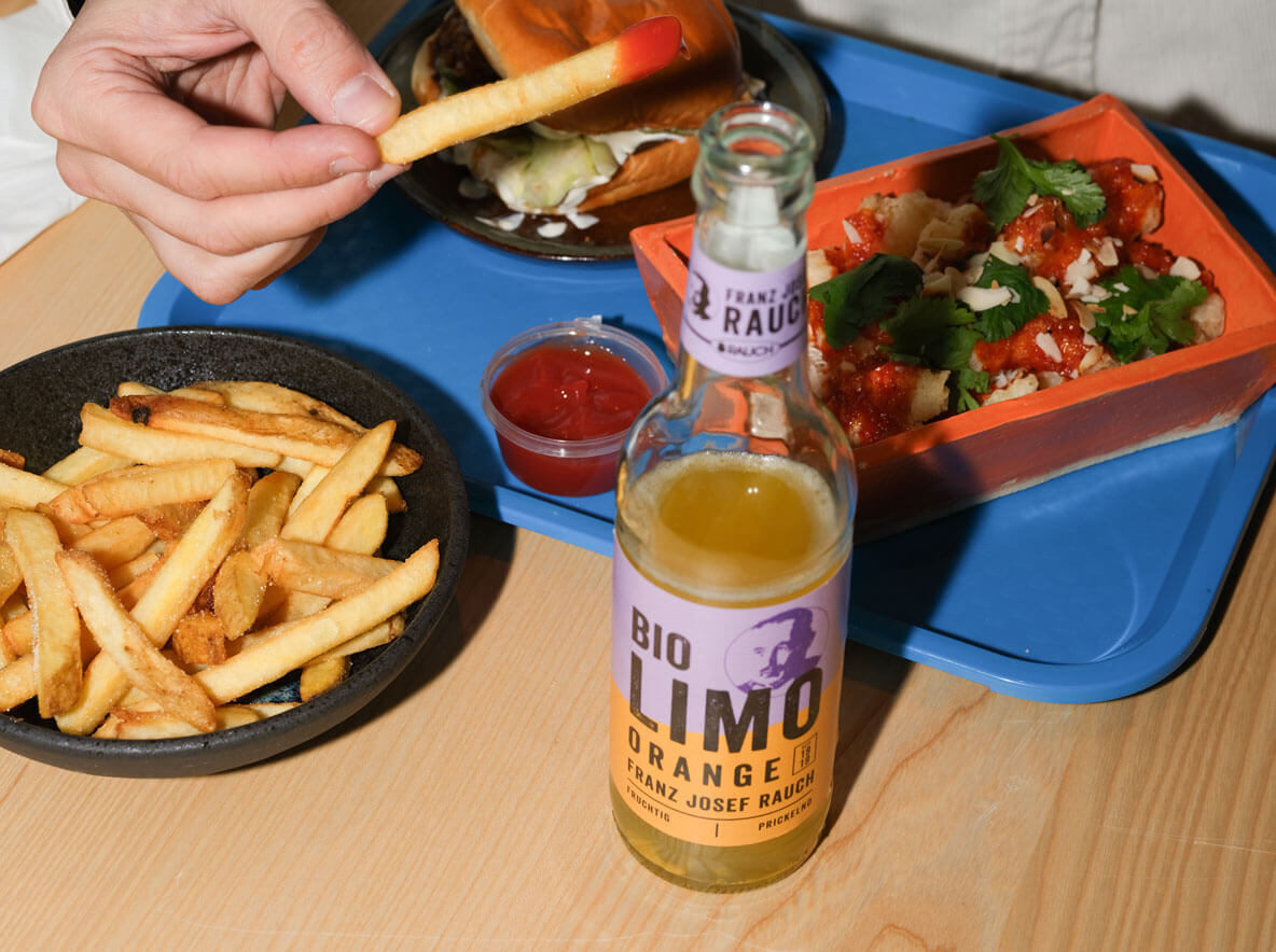 Eine Hand hält eine Pommes frites über einem Tisch mit einer Flasche Rauch Bio Orangenlimonade, einer Schüssel mit Pommes frites, Ketchup und einem Tablett mit einem Sandwich und einem Teller mit Kräutern.