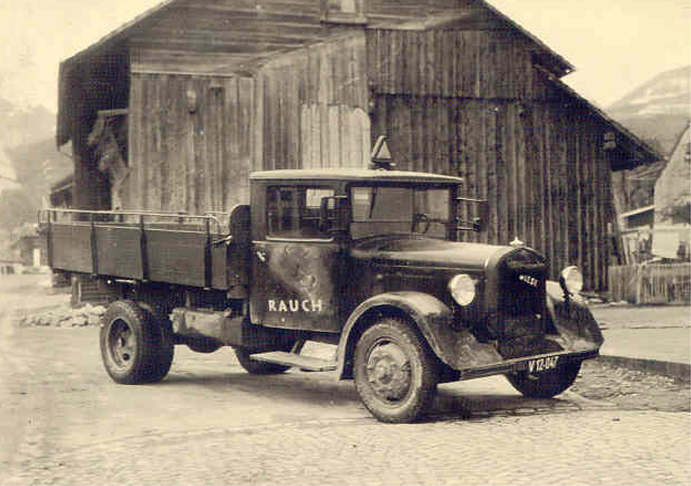 Schwarz-Weiß-Foto eines alten Pritschenwagens mit der Aufschrift "RAUCH", der vor einem Holzgebäude auf einer Kopfsteinpflasterstraße geparkt ist.