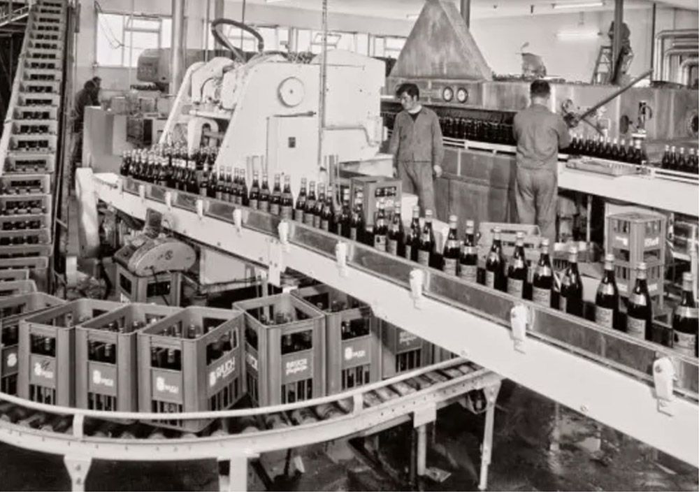 Arbeiter bedienen Maschinen an einer historischen Abfüllanlage im Rauch Werk und füllen und verpacken Glasflaschen in Kisten für den Vertrieb.