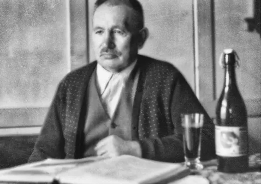 Franz Josef Rauch sitzt an einem Tisch mit einem aufgeschlagenen Buch, einem Glas und einer Flasche und blickt auf einem Schwarz-Weiß-Foto nicht in die Kamera.