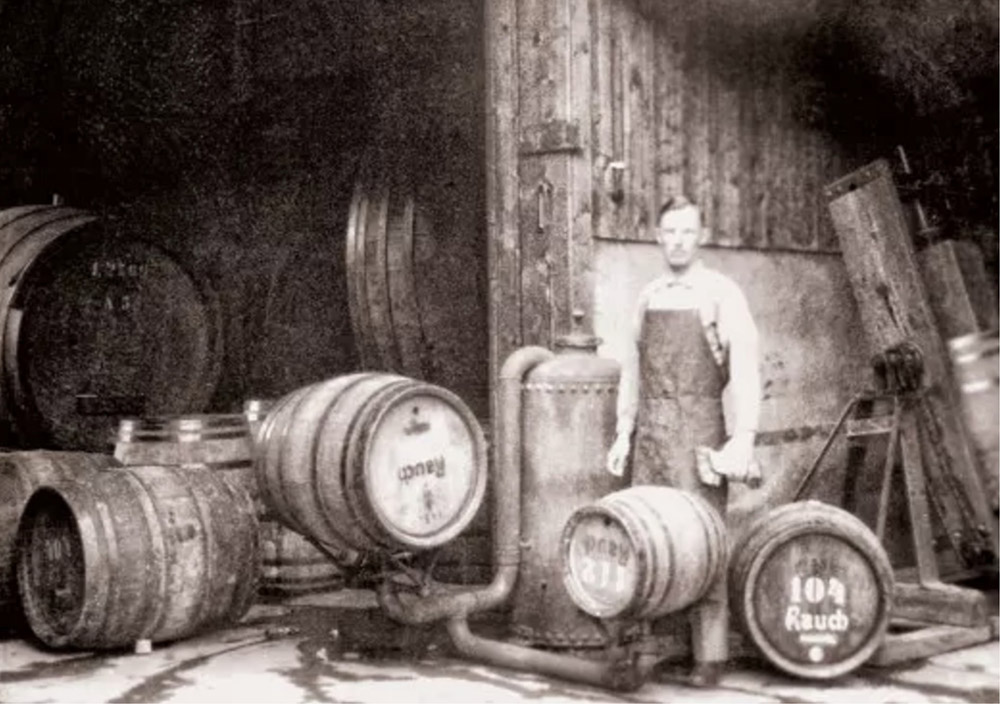 Ein Mann in einer Schürze steht zwischen Holzfässern mit der Aufschrift Rauch vor einem rustikalen Gebäude.