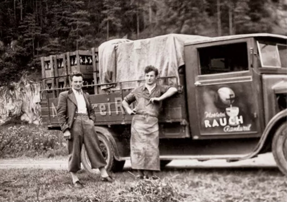Zwei Männer stehen neben einem mit Kisten und abgedeckter Ladung beladenen Rauch LKW, der im Freien in der Nähe eines Waldgebiets geparkt ist.