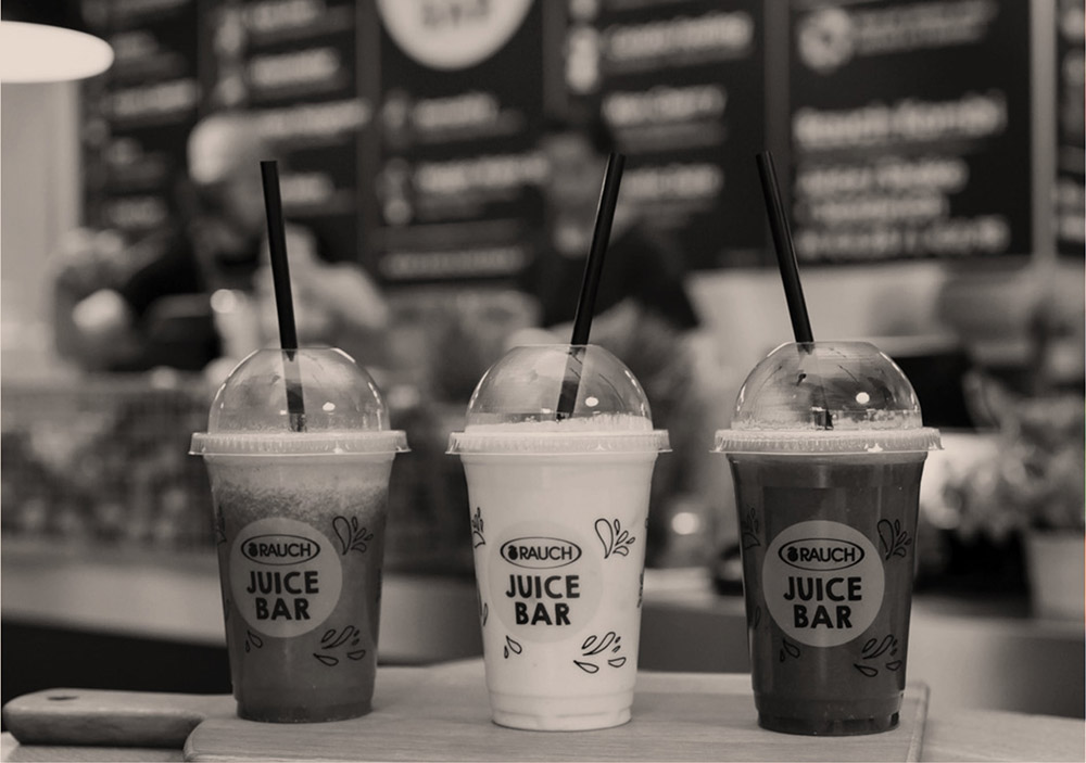 Drei Plastikbecher mit Strohhalmen und der Aufschrift "Rauch Juice Bar", die verschiedenfarbige Säfte enthalten, stehen auf einem Tresen mit einer verschwommenen Speisekarte und Menschen im Hintergrund.