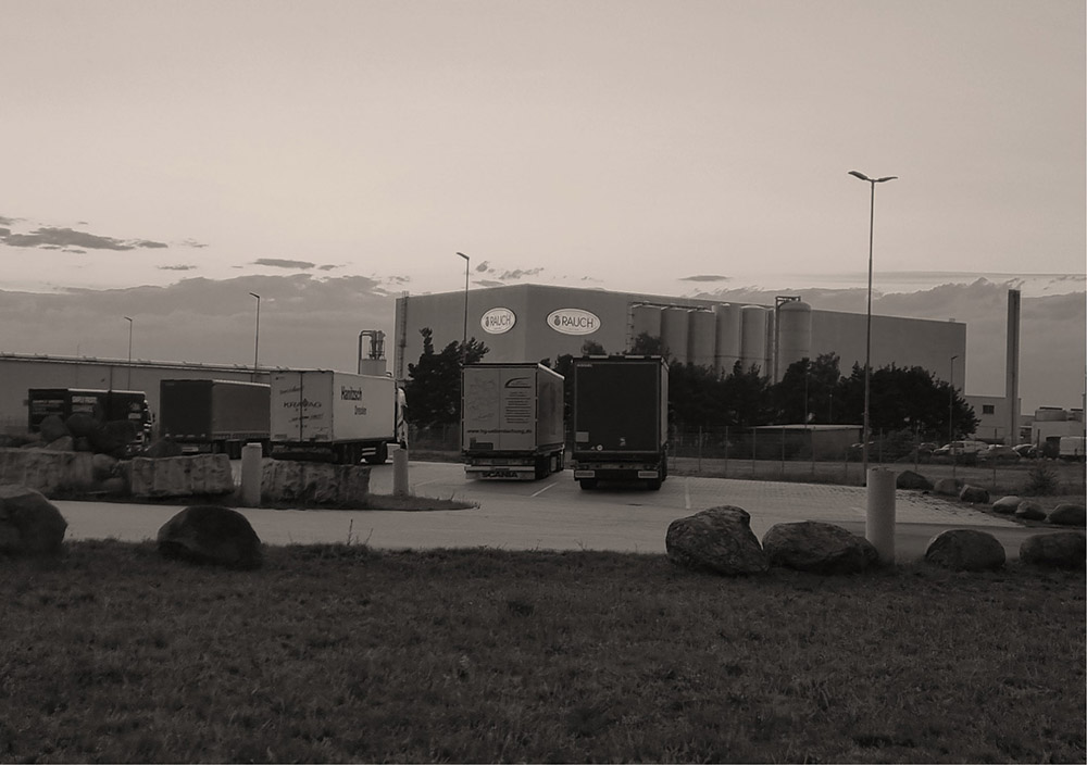 In der Nähe der Industrielagerhalle in Brandenburg mit der Aufschrift "Rauch", umgeben von Felsen und einer Grasfläche, sind bei bewölktem Himmel mehrere Lastwagen geparkt.