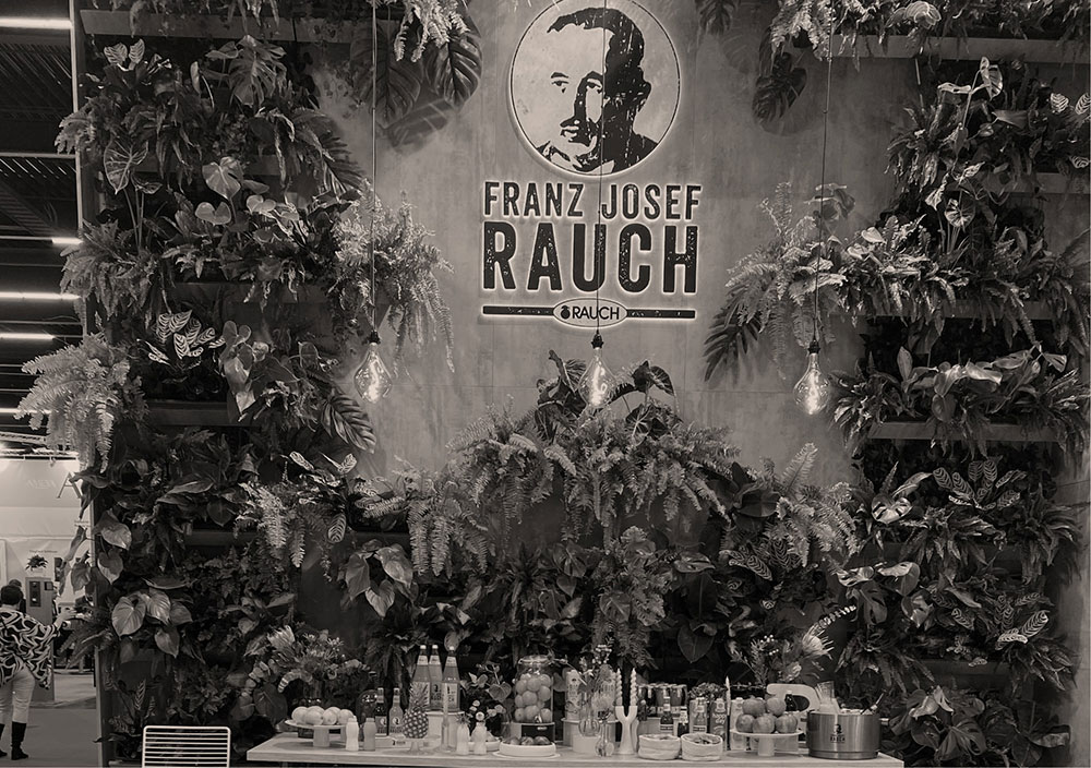 Ein Messestand mit dem Logo "Franz Josef Rauch" auf einer mit Pflanzen bedeckten Wand und verschiedenen Flaschen und Früchten auf einem Tisch davor.