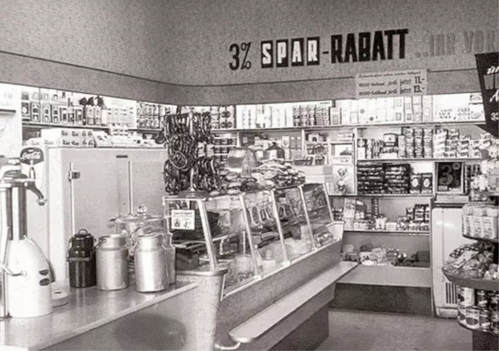Schwarz-Weiß-Foto eines kleinen Lebensmittelladens mit Warenregalen, einer Feinkosttheke und einem Schild mit der Aufschrift "3% SPAR-RABATT" über der Theke.