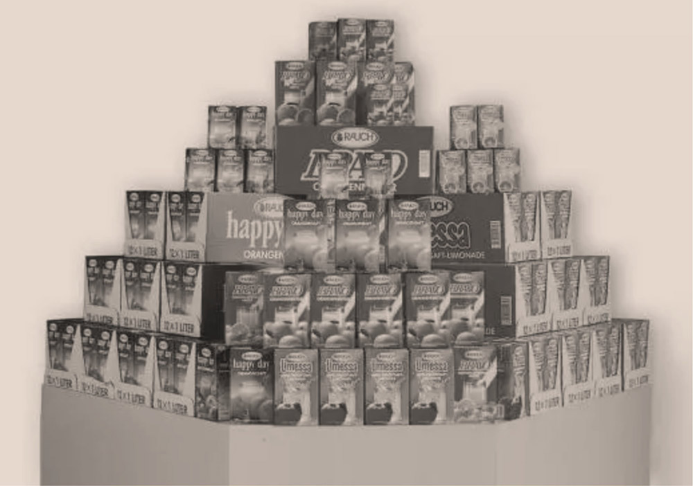 Pyramidenförmige Auslage mit verschiedenen Fruchtsaft- und Getränkekartons, die auf einer Theke gestapelt sind, darunter Marken wie Rauch Happy Day und Limessa.