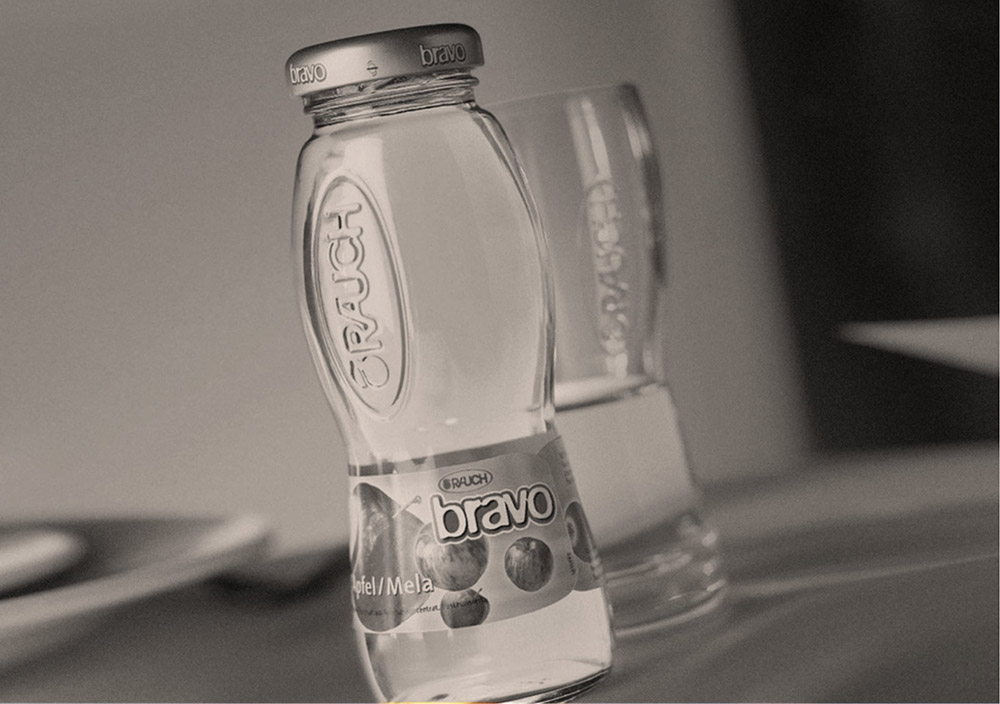 Eine Glasflasche mit der Aufschrift "Rauch Bravo" und ein Glas, die beide teilweise mit Apfelsaft gefüllt sind, stehen auf einem Tisch. Das Bild ist in schwarz-weiß gehalten.